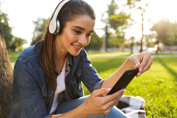 Poster - Beautiful young woman student in the park listening music with headphones using mobile phone.