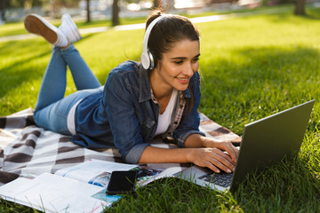 Poster - Happy woman student lies outdoors in a park using laptop computer listening music.