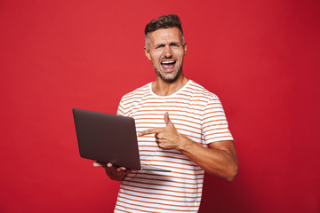 Wall Mural - Image of attractive man in striped t-shirt smiling and holding gray laptop, isolated over red background