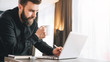 © foxyburrow - Bearded smiling businessman is standing near table, is drinking coffee, using laptop. Man works in office on computer.