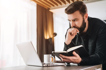 Bearded businessman is standing by computer, looking thoughtfully at laptop screen, holding notebook, watching webinar.