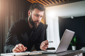 Bearded businessman is standing by computer, looking thoughtfully at laptop screen, holding pen, watching webinar.