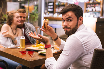 Why. Emotional man with tasty burger feeling surprised while noticing his best friends flirting