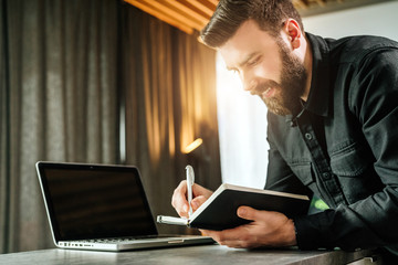 Bearded smiling businessman is standing by laptop, writing in notebook. Entrepreneur analyzes information, compares data