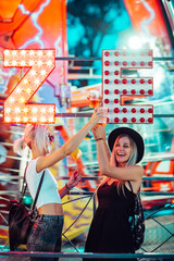 Wall Mural - Happy female friends in amusement park drinking beer. Two young women enjoying night at amusement park. 