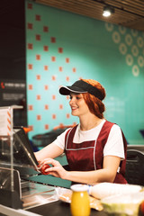 Wall Mural - Young smiling female cashier in uniform joyfully using cashbox while working in modern supermarket