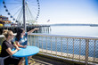 Two women looking out at the Seattle harbor on a sunny day. Sitting at a table on the waterfront near the Ferris Wheel and pier