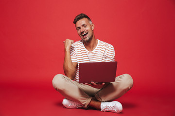 Wall Mural - Image of handsome man 30s in striped t-shirt smiling and using laptop, while sitting on floor with legs crossed isolated over red background