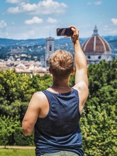 The Blond Man Takes A Photo On His Smartphone With His Back To The Camera On The Background Of A Panoramic View Of The City Of Florence, The Roof And The Cathedral Of Boboli Park