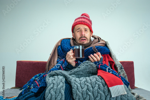 Cuadros en Lienzo Bearded sick man with flue sitting on sofa at home or studio with cup of tea covered with knitted warm clothes