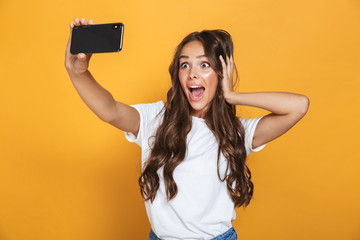 Wall Mural - Portrait of caucasian woman 20s with long hair yelling in surprise while taking selfie photo on smartphone, isolated over yellow background