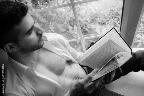 Fotografia, Obraz Man in sitting on window ledge with open shirt and pecs reading hardback book