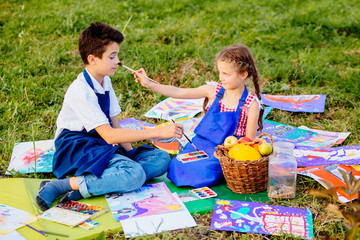 Wall Mural - Two kids brither and sister in blue aprons tickle nose each other by paint brushes, sitting outdoor in autumn time on the grass. Learning painting drawing art school concept.