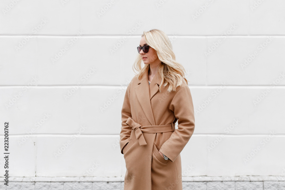 Beautiful young stylish blonde woman wearing beige coat and black sunglasses standing near white street wall. Trendy casual outfit. Street fashion. - obrazy, fototapety, plakaty 