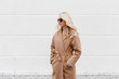 Beautiful young stylish blonde woman wearing beige coat and black sunglasses standing near white street wall. Trendy casual outfit. Street fashion.