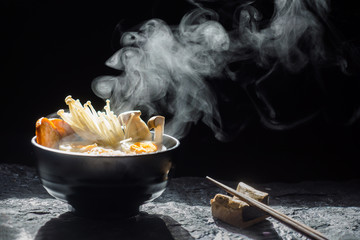 Hand uses chopsticks to tasty noodles with steam and smoke in bowl on dark background, selective focus. Asian meal on a table, junk food concept