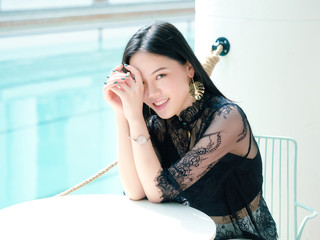 Portrait of beautiful Chinese girl in sexy black dress sitting at table and smiling at camera with blue swimming pool background in sunny afternoon.