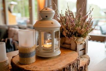 white lantern with burning candle inside it standing on festive table decorated on wooden slice and flowers on background. Decor. Wedding.Autumn still life. Toned image.Pink heather