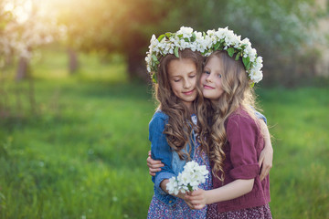 Two cute little girls friends with wreath of flowers in a blooming apple orchard