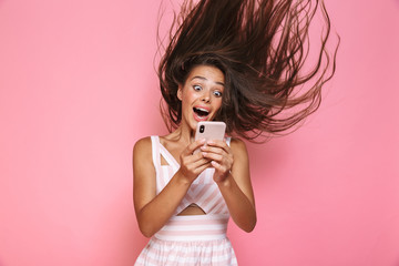 Wall Mural - Photo of caucasian woman 20s wearing dress smiling and holding mobile phone with hair shaking, isolated over pink background