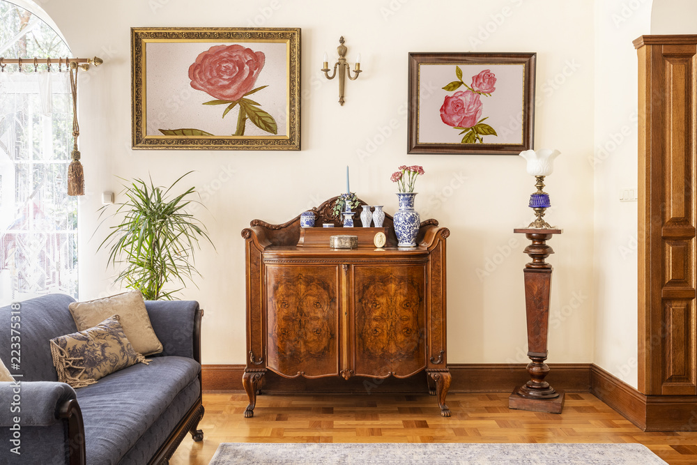 Real photo of an antique cabinet with porcelain decorations, paintings with roses and blue sofa in a living room interior - obrazy, fototapety, plakaty 