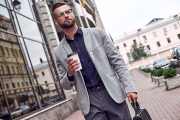 Wall Mural - Enjoying his coffee break. Handsome young man holding disposable cup and briefcase while walking outdoors pensive