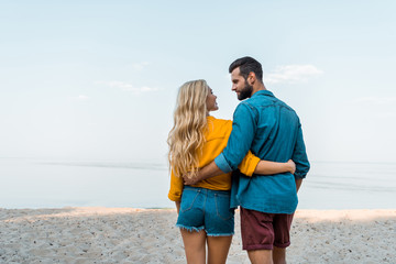 back view of couple hugging, walking and looking at each other on beach