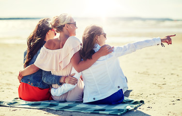 Canvas Print - summer vacation, holidays, travel and people concept - group of smiling young women in sunglasses sitting on beach blanket and pointing finger to something