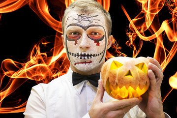 Halloween man with pumpkin against burning flame of fire background. Devil Halloween make-up. Festive scary halloween makeup. Man with pumpkin for celebration