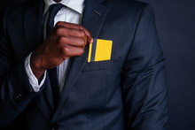 Young Successful Businessman Man In A Stylish Black Classic Suit And In Cool Glasses Holding A Yellow Plastic Credit Card In The Studio On A Dark Background. Shopping Concept