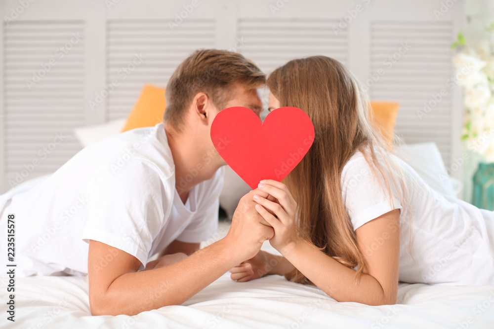 Happy young couple hiding faces behind red heart while lying on bed at home