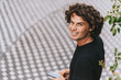 © Z A C - Rear view image of cheerful man in black t-shirt reading text message on his mobile phone on the street. Young Caucasian male with curly hair looking to the camera with copy space for text.