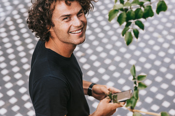 Rear overhead view of smiling man in black t-shirt typing text message on his mobile phone on the street. Young Caucasian male with curly hair looking to the camera with copy space for text.