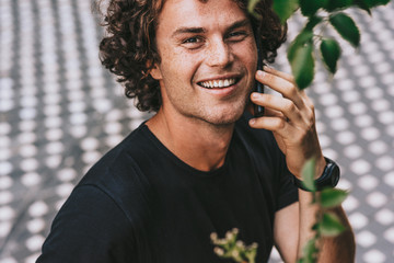 Closeup portrait of handsome young man standing outdoors talking on the mobile phone. Young man looking to the camera and making a phone call to his student friend.