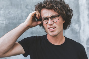 Poster - Uncertain handsome young male posing for advertisement wears black t-shirt and round glasses, isolated on gray concrete wall with copy space for your informational text. Young man feel upset. Emotion