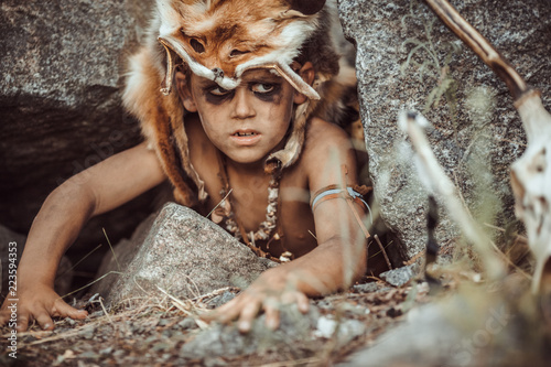 Plakat Caveman, manly boy hunting outdoors. Prehistoric tribal boy outdoors on nature. Young shaggy and dirty savage, warrior and hunter hiding in an ambush behind a stone in cave. Primitive ice age man in
