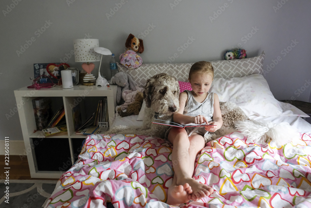 High angle view of cute girl reading book while sitting with dog on bed ...