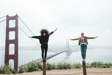 Full Length Of Playful Female Friends With Arms Outstretched Balancing On Wooden Posts Against Golden Gate Bridge During Foggy Weather