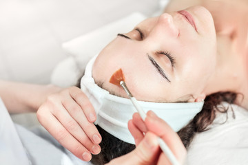 Wall Mural - The cosmetologist applies oil (or phytic acid) to the client's face in the beauty salon. A female hands of a beautician with a brush. Facial skincare. Beautiful woman on a cosmetology procedure