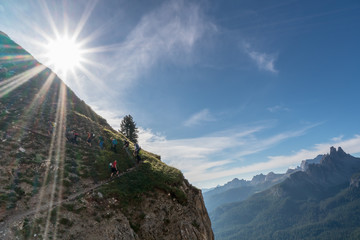 Canvas Print - large group of young hikers going up a steep hiking trail with the sun shining above them and fantastic view of mountain landscape behind