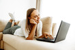 © PinkCoffee Studio - Technology. Home. Relax. Young woman is using a laptop and smiling while lying with on a couch at home