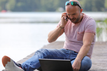 41-year-old athletic man works in Brandenburg near a lake with smartphone and laptop.