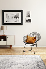 Orange pillow on a black, industrial net chair by a white wall with gallery of mock-up pictures in an elegant living room interior.