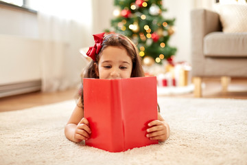 christmas, holidays and childhood concept - happy little girl lying on floor and reading book at home