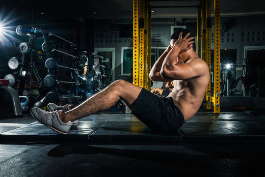 Fototapete - Muscular guy doing sit ups at gym with other people in background. Young athlete doing stomach workout in modern gym. Handsome fit man doing crunches at gym.