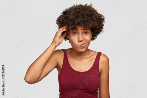 Fotografía Clueless hesitant black teenager has uncertain facial expression, curves lips, keeps hand on head, ponders on something, dressed in casual vest, isolated over white background