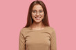 © wayhome.studio  - Horizontal view of smiling brunette young woman with pleasant appearance, wears round transparent glasses, beige sweater, models in studio against pink background. Positive emotions concept.