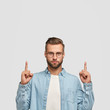 Photo of unshaven hipster dressed in casual shirt, points with both index fingers upwards. shows free space for your promotional content or advertisement. Lets go upstairs. Indoor vertical shot