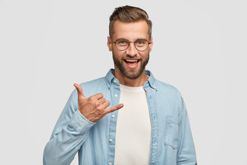 Friendly looking positive hipster with dark stubble, gestures indoor, shows shaka sign, being in high spirit, wears round spectacles for good vision, fashionable shirt, isolated on white wall