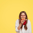 © wayhome.studio  - Pleasant looking female enjoys delicious slice of pizza, being hungry, looks with dreamy expression upwards, likes this taste, wears eyewear and white oversized shirt, stands against yellow background